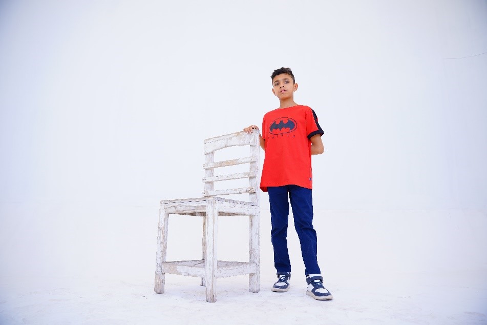 A young boy in a red Batman t-shirt and blue pants standing next to a weathered white chair against a plain background